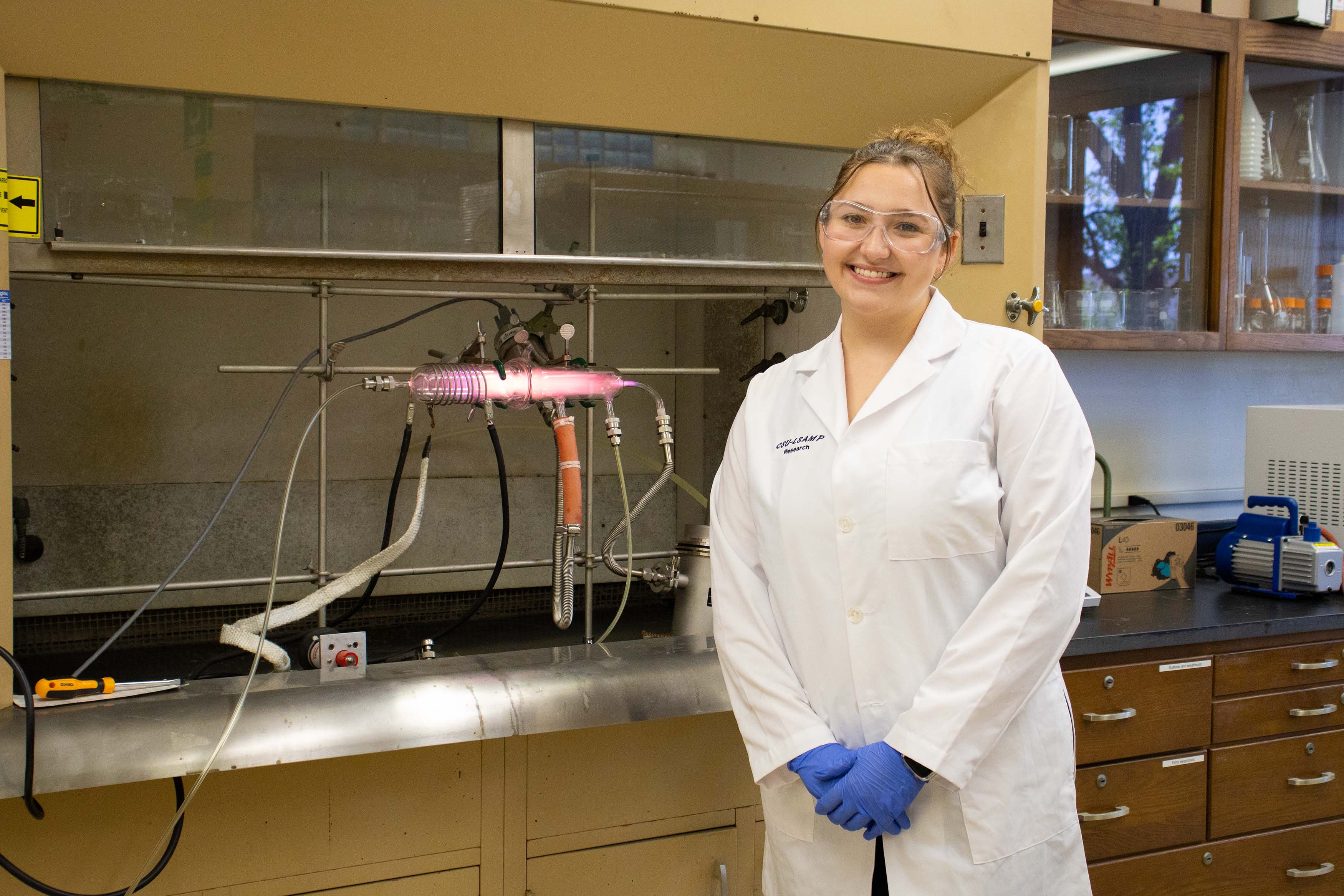 female research student in white coat standing in lab infront of a plasma reactor