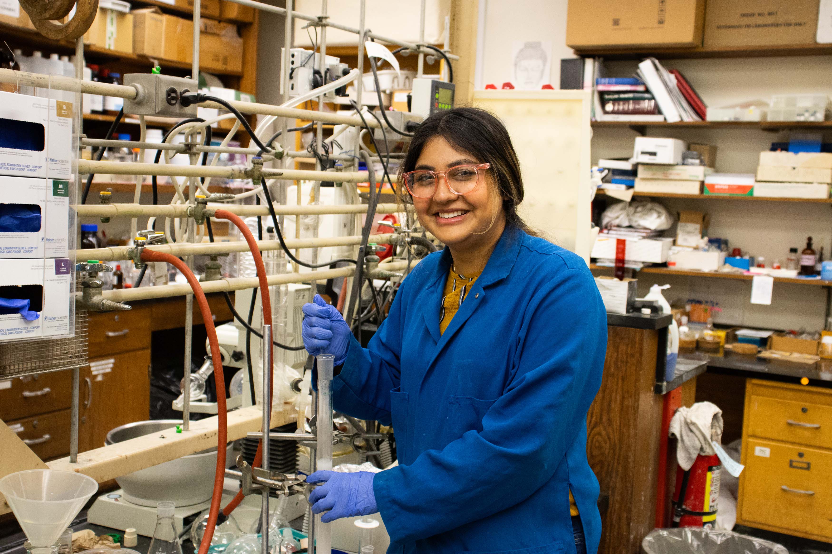 Student researcher is standing next to a bench with chemicals.