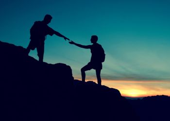 A dark blue sky up high fading to an orange sunset below in the background and two black figures of people on top of a mountain, with the people assisting each other to climb to the top.