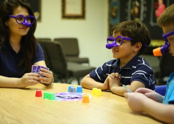 Ready to Learn – An adult and child wearing funny glasses while playing a game, with the child sitting with his arms on the table and hands folded together near his chest.