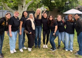 A group of people standing together, arm in arm, smiling at the camera wearing all black shirts that read “Fresno State” with a green bird raised up in the background.