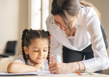 A girl sitting at a desk looking at a paper while an adult leans forward to assist the girl